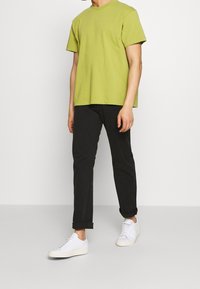 Limetkově zelené bavlněné tričko s krátkým rukávem, kombinované s černými slim-fit kalhotami a bílými teniskami s ohrnutými manžetami.