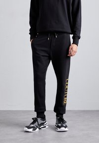 Schwarze Jogginghose aus weichem Stoff mit Kordelzug an der Taille und Seitentaschen. Goldener "COUTURE"-Schriftzug verläuft längs eines Beins. Kombiniert mit schwarz-weißen Sneakers.