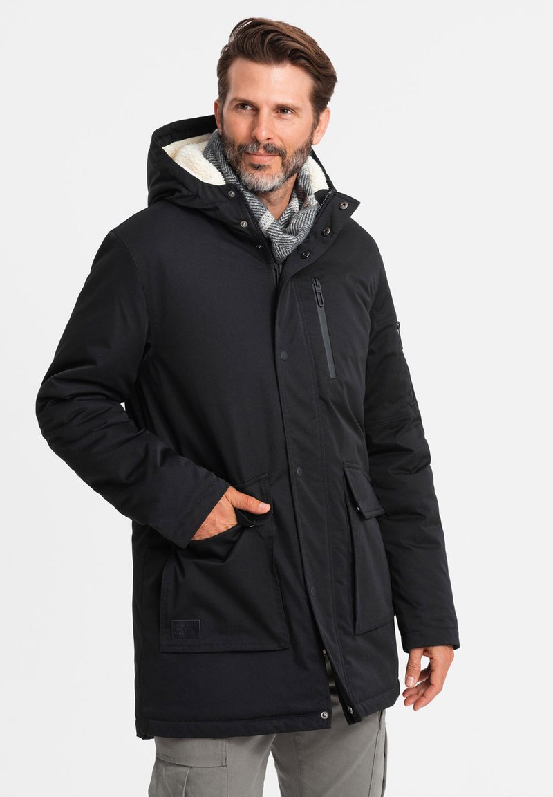 Parka nera isolata con cappuccio, tasca sul petto con zip e tasche anteriori con bottoni. Presenta un caldo rivestimento in faux shearling.