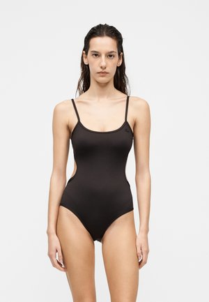 Femme aux cheveux mouillés portant un maillot de bain une pièce noir avec des bretelles fines et des côtés ouverts, debout devant un fond clair uni.