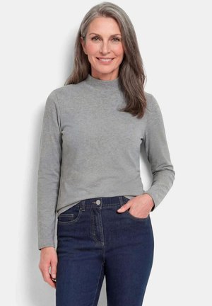 Femme aux longs cheveux gris portant une chemise grise à manches longues et un jean bleu foncé, souriante, une main dans sa poche.