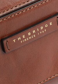 Portafoglio in pelle marrone con superficie testurizzata, dettagli cuciti e un'etichetta nera con scritte dorate che riportano "THE BRIDGE FIRENZE 1969."