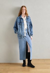 Joven mujer con cabello largo rojo que lleva una camisa blanca, una chaqueta de mezclilla azul oversized, una falda de mezclilla larga con apertura frontal y botas negras.