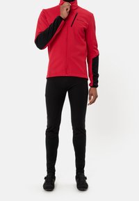 Veste en polaire rouge avec des accents noirs et une poche poitrine zippée, associée à un pantalon noir près du corps et des chaussures de cyclisme, sur fond blanc.