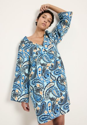 Woman wearing loose, long-sleeve dress with blue, black, and peach paisley pattern, posing with one arm raised by her head.