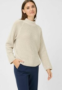 Beige gestrickter Pullover mit hohem Kragen, der ein strukturiertes Muster und eine lockere Passform aufweist, kombiniert mit navyblauen Hosen.