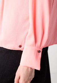 Manche de blouse rose clair avec poignet boutonné sur le bras d'une personne, associée à un pantalon noir texturé.