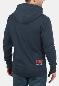 Dark gray hoodie featuring a back pocket, a drawstring hood, and a bold red logo with "Since 1993" near the hem. Soft fabric texture.