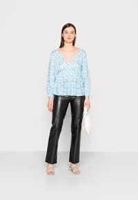 Blusa floral azul clara com decote em V, mangas longas, cintura franzida, combinada com calças de couro pretas e sandálias azuis claras. Segurando uma bolsa branca.
