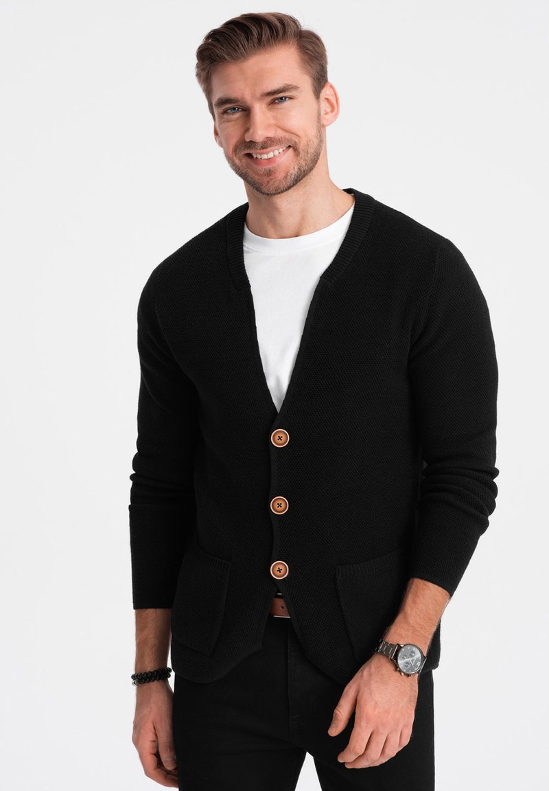 Černý pletený kardigan s véčkovým výstřihem, se dřevěnými knoflíky, dvěma předními kapsami a žebrovanými manžetami. Nositelný přes bílé tričko.