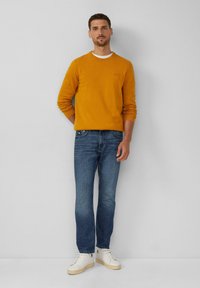 Mosterdgele gebreide trui met ronde hals, gecombineerd met donkerblauwe denim jeans en witte sneakers voor een casual uitstraling.