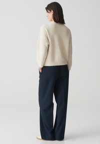 Beige strukturiertes Strickpullover mit einem lässigen Schnitt, kombiniert mit marineblauen weit geschnittenen Hosen und weißen Sneakers, von hinten betrachtet.