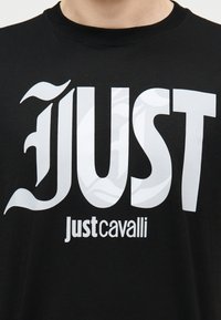 Μαύρο μπλουζάκι με μεγάλη λευκή στιλιζαρισμένη λέξη «JUST» και μικρότερο κείμενο μάρκας «just cavalli» κάτω από αυτήν στο στήθος.
