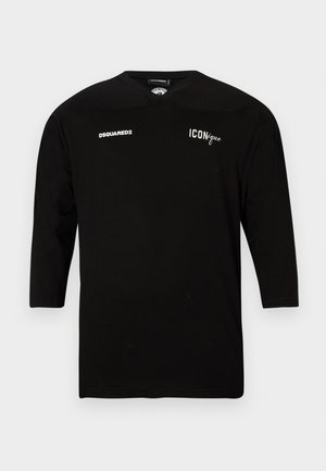 Jeune homme aux cheveux bouclés portant un t-shirt noir à manches longues avec des logos, un jean bleu déchiré et des baskets noires, debout les mains dans le dos.