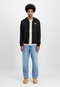 Sudadera negra con cremallera, cordones y logo, llevada sobre una camiseta beige; combinada con jeans de pierna ancha de color azul claro y calzado en tonos marrones.