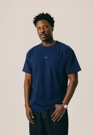 Mann mit kurzen lockigen Haaren, der ein dunkelblaues T-Shirt, dunkle Jeans, eine Armbanduhr und eine goldene Kette trägt und vor einem schlichten beigefarbenen Hintergrund steht.