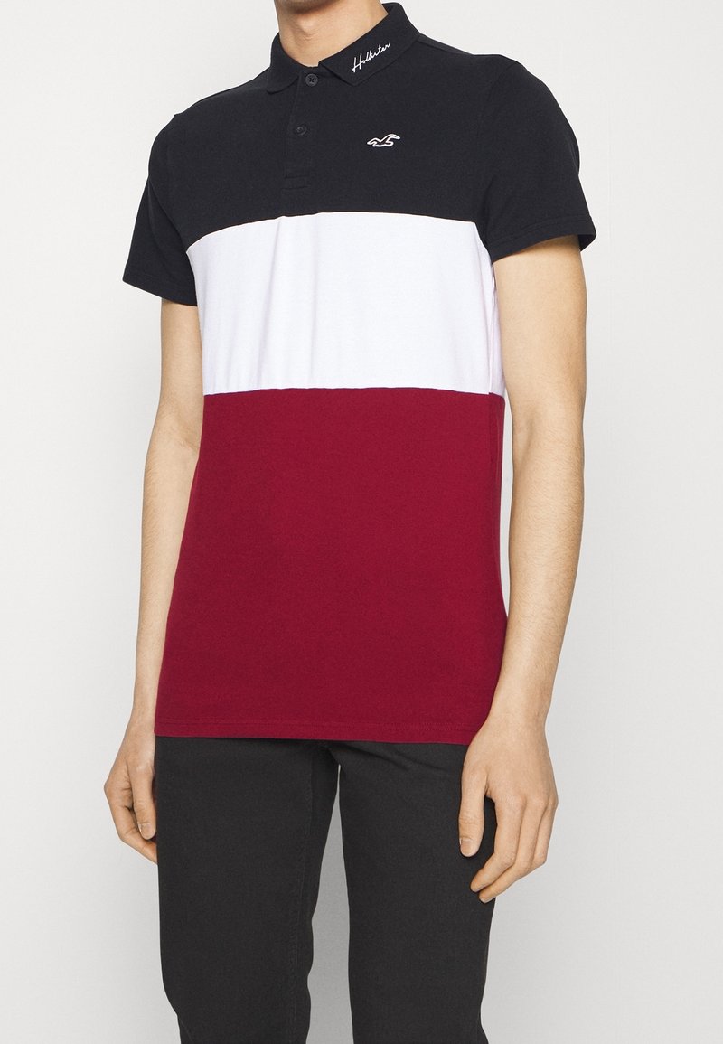 Homme portant un polo à manches courtes avec col noir, partie supérieure noire, milieu blanc et partie inférieure bordeaux, associé à un pantalon noir.
