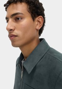 Giacca in suede verde scuro con chiusura a zip, colletto ripiegato e tasca laterale. Il modello presenta riccioli leggeri e un piccolo orecchino a cerchio in argento.