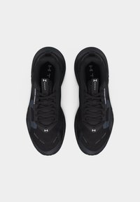 Ζευγάρι μαύρα αθλητικά παπούτσια Under Armour με κορδόνια, εμφανές λογότυπο και υφασμένες σόλες, που φαίνονται από πάνω σε λευκό φόντο.