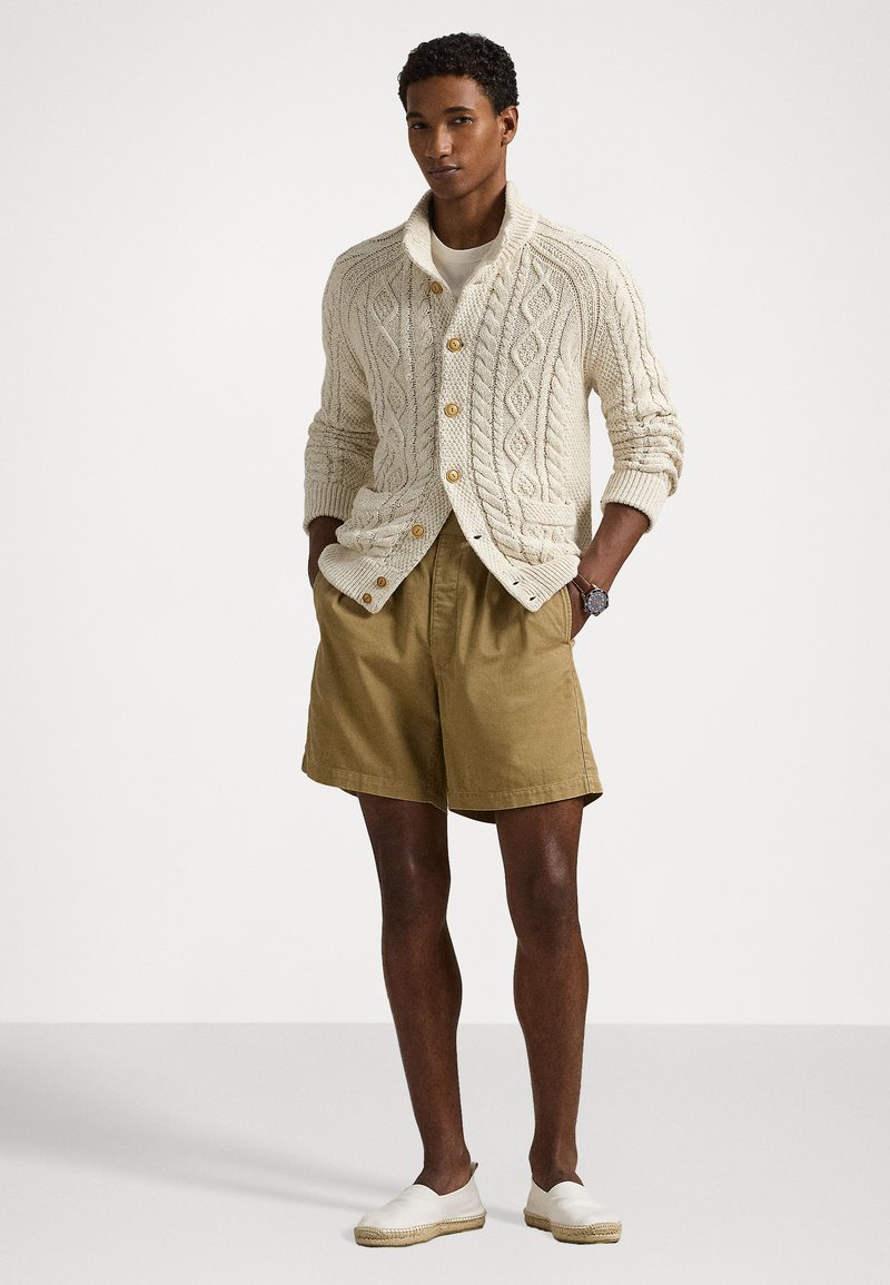 Cardigan a maglia a trecce crema con bottoni in legno, indossato sopra una maglietta beige, abbinato a pantaloni corti khaki e scarpe slip-on bianche.