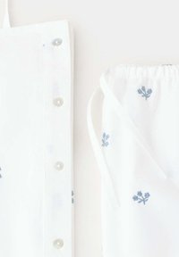 Tissu blanc avec une petite broderie florale bleu clair, présentant des boutons sur le côté gauche et un cordon froncé sur le côté droit.