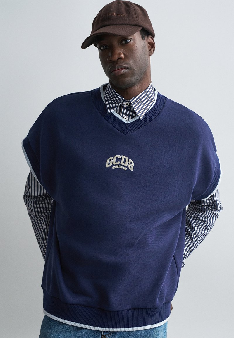 Sweater oversized azul marinho com decote em V, apresentando um logótipo bordado em branco. Camisa de colarinho riscada em azul e branco visível por baixo do sweater.