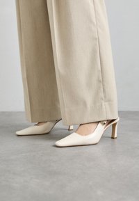 Minkštos baltos odos slingback kulniukai, smailas pirštas, akcentuoti mažu auksiniu sagtimi, derinami su šviesiai rudos spalvos plačiomis kelnėmis.