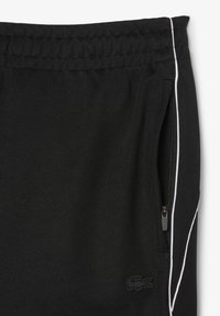 Zwarte sportbroek van een gestructureerde stof, met een zijzak met rits en witte piping langs de naden. Lacoste-logo op de zoom.