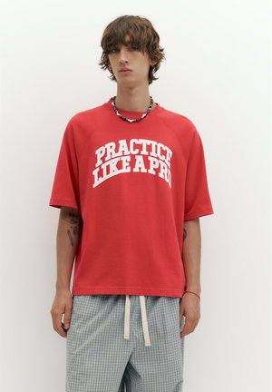 Jeune personne aux cheveux bruns courts porte un t-shirt rouge oversize avec le texte "PRACTICE LIKE A PRO", un pantalon à carreaux et un collier de perles.