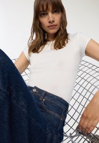 Biała dopasowana koszulka z krótkim rękawem w połączeniu z ciemnoniebieskimi dżinsami. Dżinsy mają pomarańczowe przeszycia i srebrny guzik. Tło z metalowej kraty krzesła.