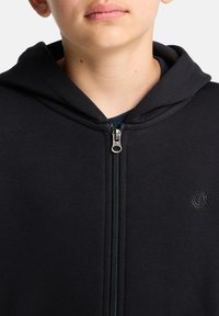 Felpa nera con zip realizzata in tessuto testurizzato. Presenta una zip frontale, un cappuccio e un dettaglio del logo sul lato sinistro.