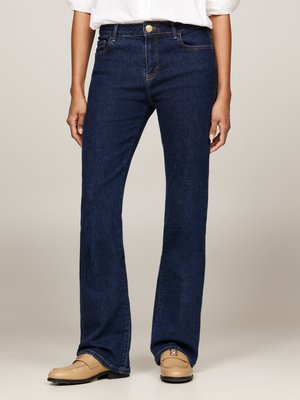 Jeans de mezclilla oscura en corte acampanado con cierre de botón dorado, diseño de cinco bolsillos, costuras en tono y una textura suave. Llevados con zapatos beige.