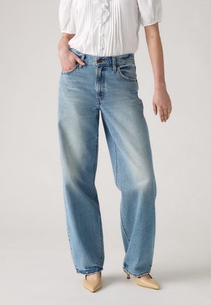 Femme portant un jean large bleu clair, un chemisier blanc à manches courtes avec des volants, des talons beige à bouts pointus, debout avec une main dans la poche.