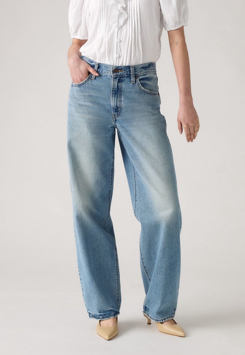 Femme portant un jean large bleu clair, un chemisier blanc à manches courtes avec des volants, des talons beige à bouts pointus, debout avec une main dans la poche.