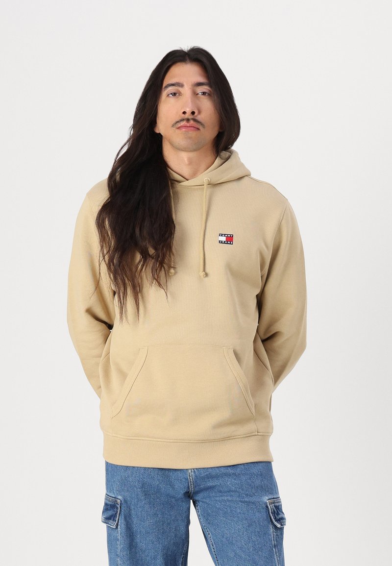Tan hoodie made of ribbed fabric, featuring a front pouch pocket, drawstring hood, and a small logo patch in red, white, and blue.