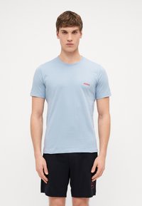 Light blue cotton T-shirt with a crew neck and short sleeves, featuring a small red "HUGO" logo on the left chest. Worn with dark shorts.