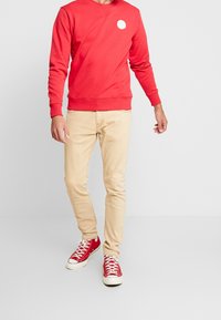 Red sweatshirt with a round logo on the chest, beige fitted jeans, and red sneakers with white accents. Simple design, casual look.