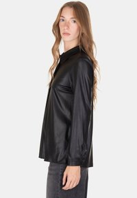 Camicia nera in pelle a maniche lunghe con colletto, caratterizzata da una texture liscia e polsini con bottoni, indossata sopra jeans scuri in denim.