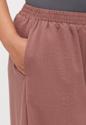 Personne portant un pantalon mauve à taille élastique, main posée négligemment dans la poche latérale, montrant la texture du tissu et les détails de couture.