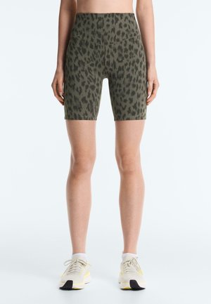 Kobieta w zielonych, wysokowzwyżających szortach rowerowych w panterkę i białych trampkach z żółtymi akcentami, stojąca na białym tle.