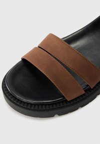 Sandale à glisser en cuir brun avec deux larges bretelles, semelle texturée noire et bout arrondi. Présente des détails cousus et une semelle intérieure contournée.