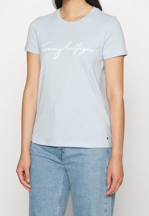 Vrouw die een lichtblauw Tommy Hilfiger T-shirt draagt met wit scriptlogo en lichtblauwe spijkerbroek, staand tegen een effen witte achtergrond.