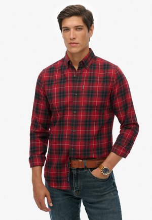 Homme portant une chemise à carreaux rouge et noir à boutons avec manches retroussées, jean bleu, ceinture marron et montre-bracelet, debout avec une main dans la poche.