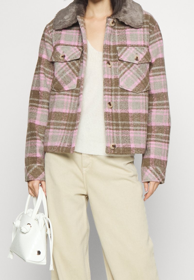Personne portant une veste à carreaux rose et marron avec de gros boutons, un pantalon beige et tenant un petit sac à main blanc avec des franges.