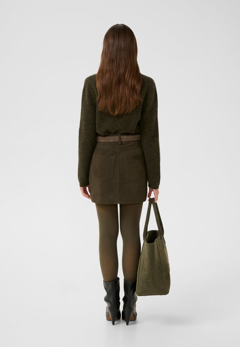 Grüner Pullover, brauer Wildleder-Minirock mit Taschen, olivgrüne Strumpfhosen, schwarze Stiefeletten mit Absatz, hält eine olivgrüne Tragetasche.
