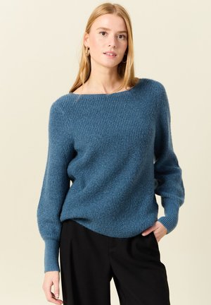 Blauer gestrickter Pullover mit geripptem Muster, weitem Ausschnitt und langen Ärmeln. Kombiniert mit schwarzen Hosen, zeigt einen lässigen Stil.