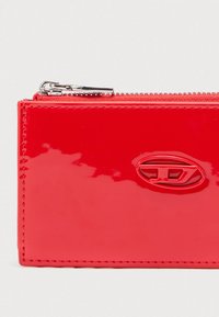 Πορτοφόλι από κόκκινο δερμάτινο λουστρίνι με φερμουάρ και εμφανές ανάγλυφο λογότυπο. Ομαλή υφή με ορατές ραφές.