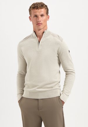 Jonge man in een beige half-zip trui en een bruine broek, staand met zijn handen in zijn zakken tegen een effen lichte achtergrond.