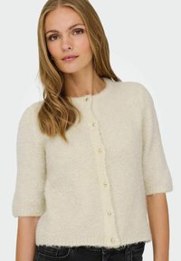 Jeune femme portant un cardigan en peluche couleur crème avec des boutons dorés et des manches trois-quarts, associé à un pantalon sombre.