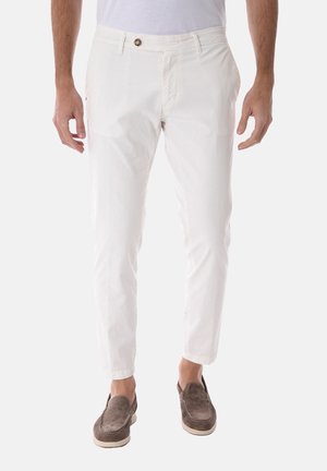 Uomo che indossa pantaloni aderenti bianchi con chiusura con bottone e zip, abbinati a scarpe slip-on in suede marrone.